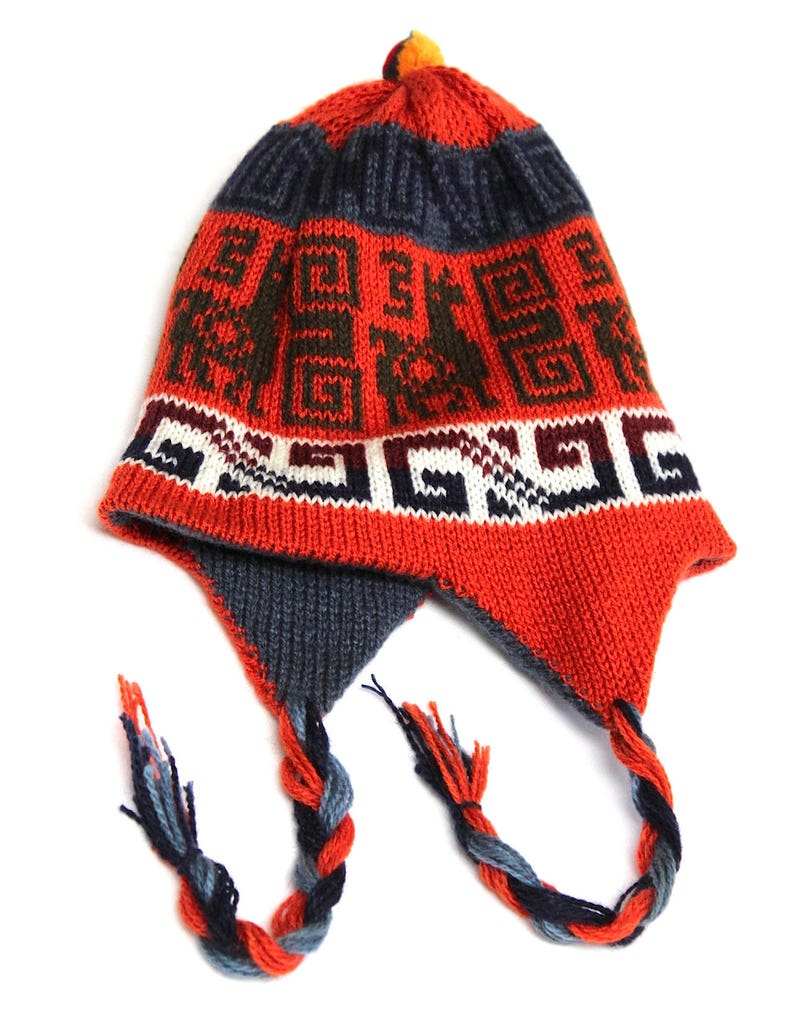 May include: A knitted hat with ear flaps, featuring a vibrant red base with geometric patterns in dark blue and brown. The hat has a braided tassel and a small orange pom-pom on top. The ear flaps are dark blue and the tassels are a mix of blue, orange, and grey.