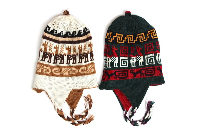 May include: Two knitted winter hats with ear flaps. One is cream-colored with brown llama patterns and the other is dark green with red and white geometric designs. Both have braided ties.