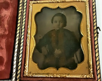 Pode incluir: Um retrato de daguerreótipo vintage de uma pessoa jovem vestindo uma jaqueta escura e uma camisa clara. O retrato é emoldurado em uma moldura dourada com um passe-partout de veludo vermelho.