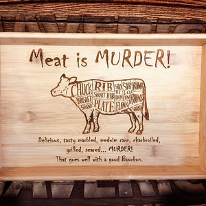 May include: A wooden serving tray with a cow silhouette and the text "Meat is MURDER!"  The cow is divided into different cuts of meat, including chuck, rib, short rib, brisket, plate, flank, and sirloin. The text below the cow reads "Delicious, tasty marbled, medium rare, charbroiled, grilled, seared... MURDER! That goes well with a good Bourbon."