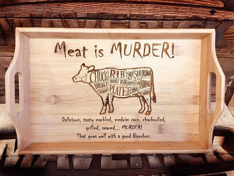 May include: A wooden serving tray with a cow silhouette and the text "Meat is MURDER!"  The cow is divided into different cuts of meat, including chuck, rib, short rib, brisket, plate, flank, and sirloin. The text below the cow reads "Delicious, tasty marbled, medium rare, charbroiled, grilled, seared... MURDER! That goes well with a good Bourbon."