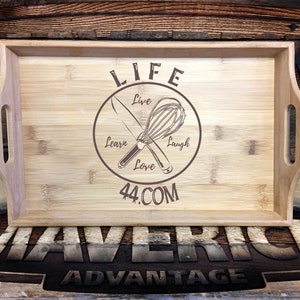 May include: A wooden serving tray with a brown circle design featuring a whisk and knife crossed over each other. The circle has the words "LIFE" at the top, "Live", "Learn", "Love", and "Laugh" around the circle, and "44.COM" at the bottom. The tray has two handles on the sides and the words "MAVERICK ADVANTAGE" are printed on the surface below the tray.