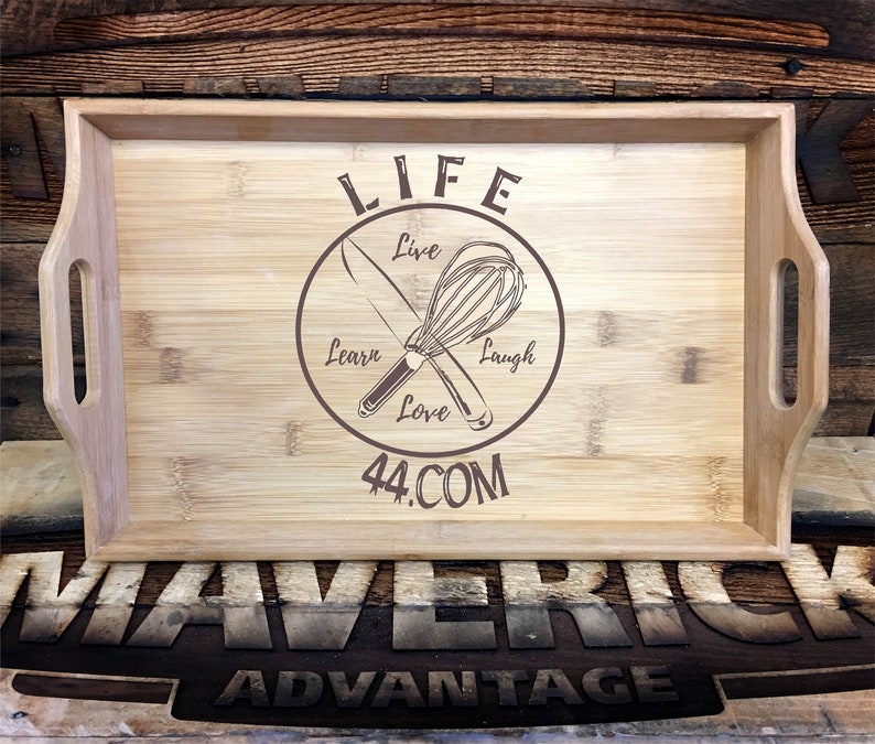 May include: A wooden serving tray with a brown circle design featuring a whisk and knife crossed over each other. The circle has the words "LIFE" at the top, "Live", "Learn", "Love", and "Laugh" around the circle, and "44.COM" at the bottom. The tray has two handles on the sides and the words "MAVERICK ADVANTAGE" are printed on the surface below the tray.