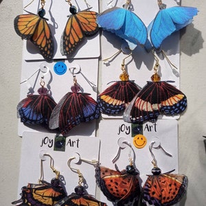 May include: Four pairs of butterfly-shaped earrings on white cards with the text "Joy Art". The earrings are made of fabric and have gold-colored hooks. The butterflies are in various colors, including orange, blue, and black.