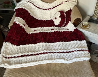 May include: A chunky knit blanket in a bold red and white striped pattern, draped over a beige armchair. A matching round pillow rests on the blanket. The blanket is made of thick, soft yarn.