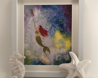 May include: A framed painting of a mermaid with red hair swimming upwards towards a small seahorse. The background is a colorful abstract design of blues, purples, and greens. The frame is white and there are two white seahorse and starfish figurines on the table below the frame.