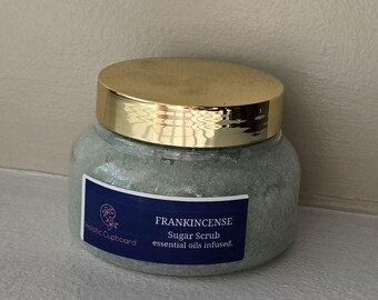 May include: A clear glass jar with a gold lid, filled with a light green sugar scrub. A blue label reads "FRANKINCENSE Sugar Scrub essential oils infused." The jar is from Holistic Cupboard.