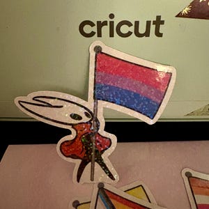May include: A sticker featuring a character holding a bisexual pride flag with red, purple, and blue stripes. The word "cricut" is printed in black on a light green background. Other pride flag stickers are visible.