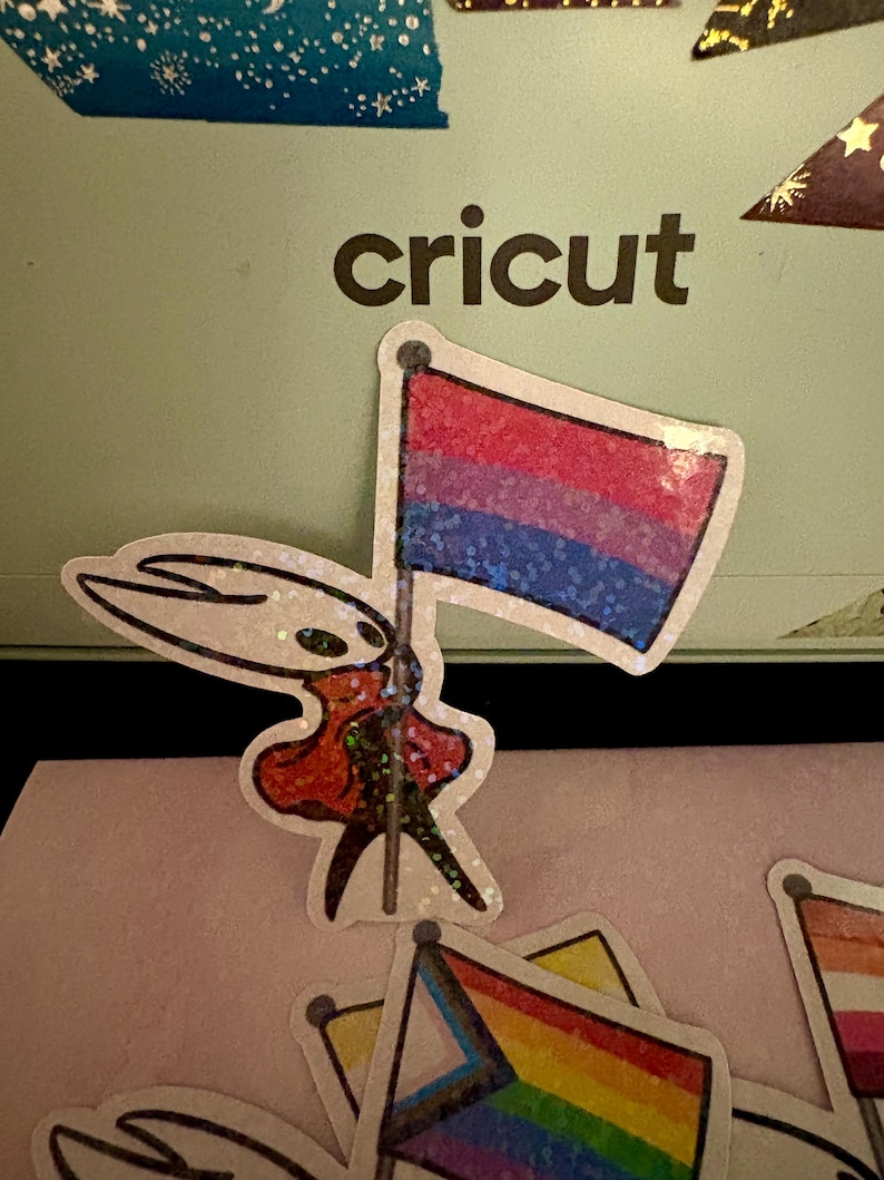 May include: A sticker featuring a character holding a bisexual pride flag with red, purple, and blue stripes. The word "cricut" is printed in black on a light green background. Other pride flag stickers are visible.