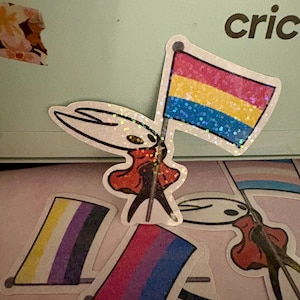 May include: A collection of stickers featuring a character holding various pride flags. The flags include the pansexual, non-binary, bisexual, and transgender pride flags. The stickers have a glittery finish and are on a light pink surface.