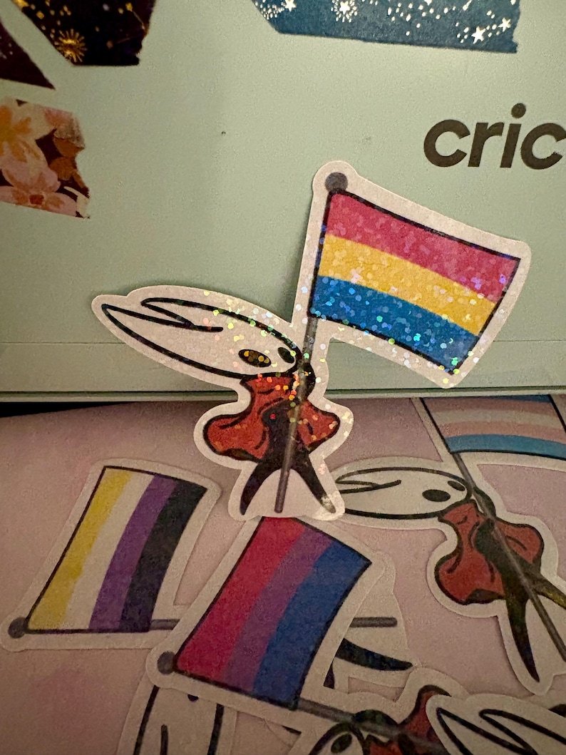 May include: A collection of stickers featuring a character holding various pride flags. The flags include the pansexual, non-binary, bisexual, and transgender pride flags. The stickers have a glittery finish and are on a light pink surface.
