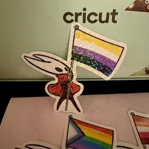 May include: A collection of stickers featuring a cartoon character holding various pride flags. The flags include the non-binary flag, the progress pride flag, and a lesbian pride flag. The stickers have a glittery finish and are set against a light green background with the word "cricut".