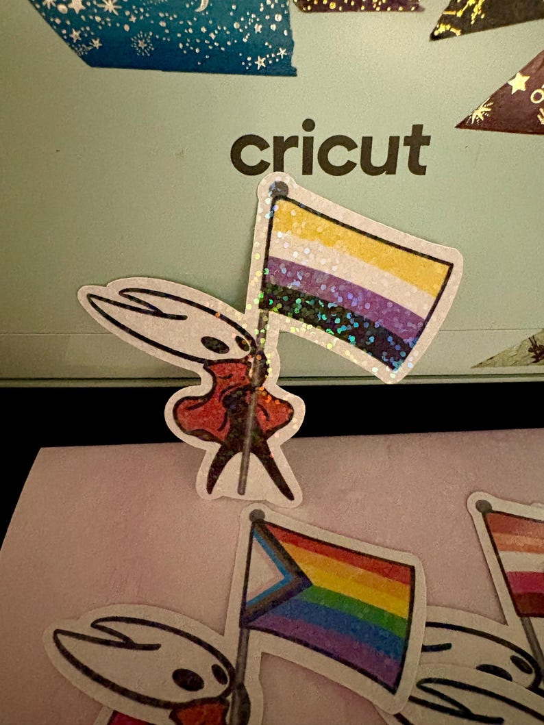 May include: A collection of stickers featuring a cartoon character holding various pride flags. The flags include the non-binary flag, the progress pride flag, and a lesbian pride flag. The stickers have a glittery finish and are set against a light green background with the word "cricut".