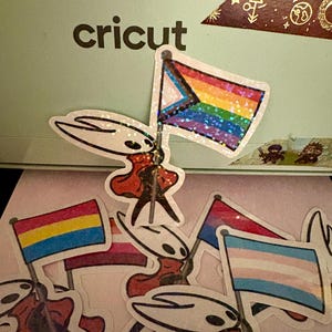 May include: A collection of stickers featuring a character holding various Pride flags. The flags include the Progress Pride flag, the Pansexual Pride flag, the Lesbian Pride flag, and the Transgender Pride flag. The word "cricut" is visible.