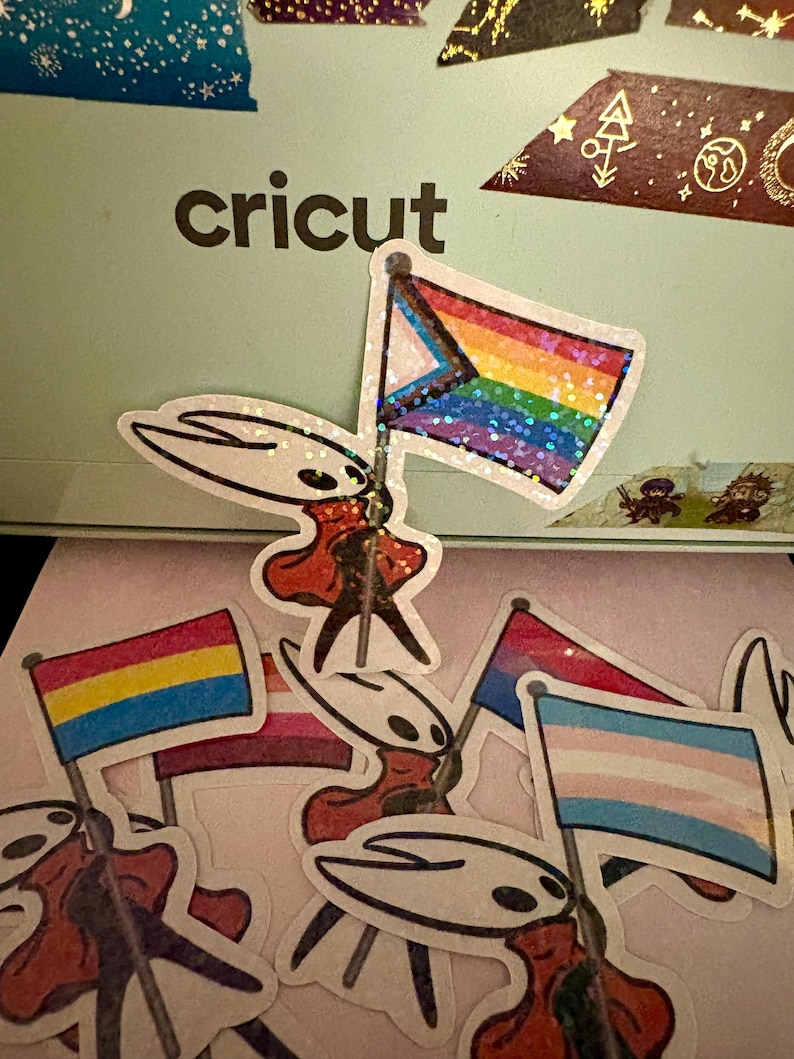 May include: A collection of stickers featuring a character holding various Pride flags. The flags include the Progress Pride flag, the Pansexual Pride flag, the Lesbian Pride flag, and the Transgender Pride flag. The word "cricut" is visible.