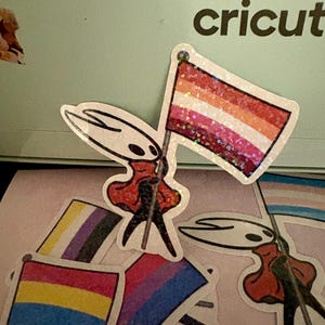 May include: A collection of stickers featuring a character holding various pride flags. The flags represent lesbian, bisexual, pansexual, non-binary, and transgender identities. The stickers have a glittery finish and are on a light pink surface.
