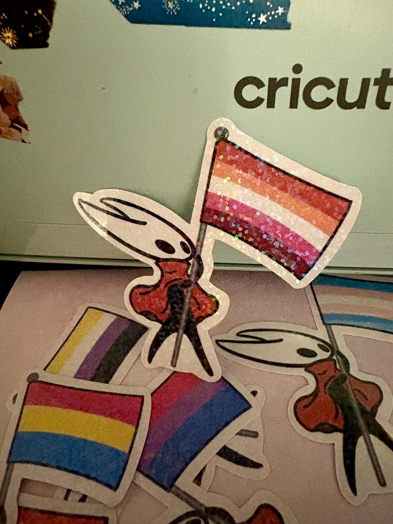 May include: A collection of stickers featuring a character holding various pride flags. The flags represent lesbian, bisexual, pansexual, non-binary, and transgender identities. The stickers have a glittery finish and are on a light pink surface.