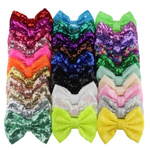 May include: A collection of 15 colorful sequined hair bows in various colors, including green, blue, pink, purple, and yellow. The bows are arranged in a row, with the largest bow at the center.