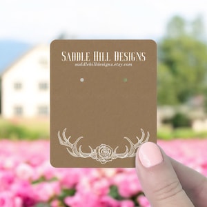 May include: Brown card stock earring display card with white antlers and a rose design. The text "Saddle Hill Designs" and the website "saddlehilldesigns.etsy.com" are printed on the card.