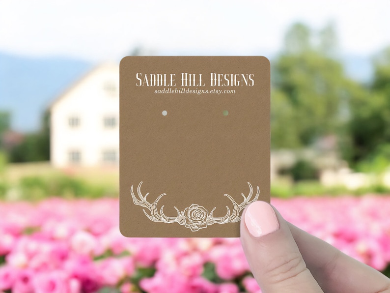 May include: Brown card stock earring display card with white antlers and a rose design. The text "Saddle Hill Designs" and the website "saddlehilldesigns.etsy.com" are printed on the card.