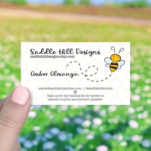 May include: White business card with black text that reads "Saddle Hill Designs" and "saddlehilldesigns.etsy.com". Below the text is a cartoon bee with black and yellow stripes flying over a dotted line. The text "Amber Almanza" is below the bee. The text "amber@saddlehillstudios.com | @saddlehillstudios" is below the name. The text "Sign up for my mailing list for access to special coupons and product updates!" is below the contact information.
