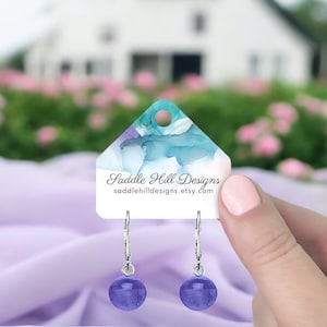 May include: A pair of silver hoop earrings with a purple glass bead dangle. The earrings are displayed on a white card with the text "Saddle Hill Designs" and "saddlehilldesigns.etsy.com".