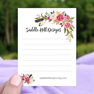 May include: A white card with a floral design in shades of pink, purple, and green. The card has the text "Saddle Hill Designs" and the website address "saddlehilldesigns.etsy.com".