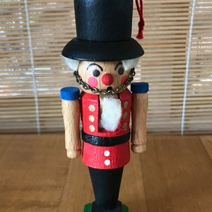 May include: A wooden nutcracker figurine with a black hat, red and white uniform, and a gold chain. The nutcracker is standing on a green base.