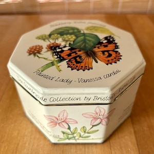 May include: A white octagonal tin with a gold trim. The tin features a butterfly with orange and black wings, green leaves, and pink flowers. The text on the tin reads "Painted Lady - Vanessa cardui" and "The Countryside Collection by Bristows - Rural Life".