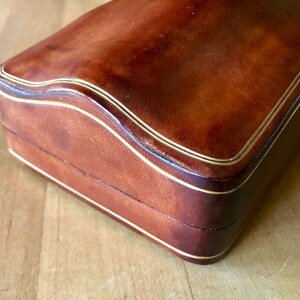 May include: A brown leather box with a gold trim. The box has a curved top and a hinged lid.