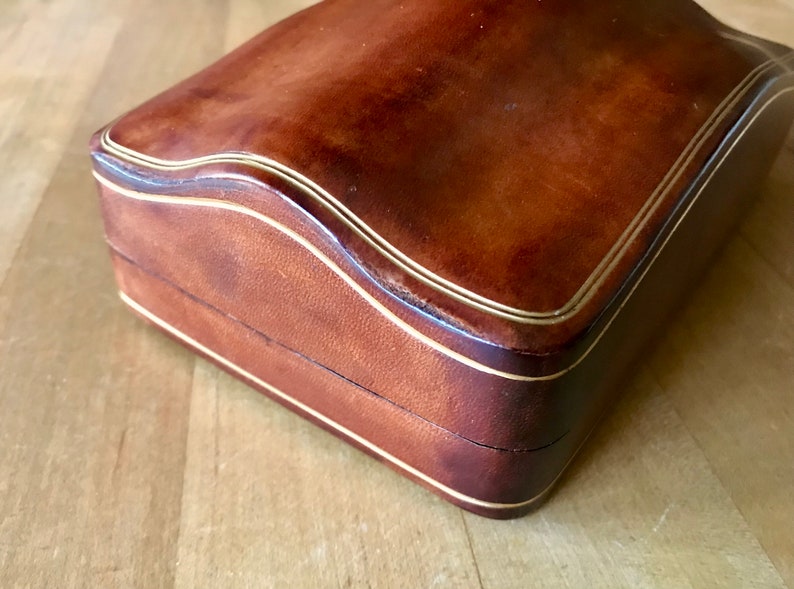 May include: A brown leather box with a gold trim. The box has a curved top and a hinged lid.