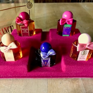 May include: A set of five miniature perfume bottles in a red velvet tray. The bottles are all different colors and have different labels. The labels include the names "White Shoulders", "Passion", "Magnolia", "Bamboo", and "Enchantment".