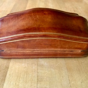 May include: A brown leather box with a gold trim. The box has a curved top and a hinged lid.