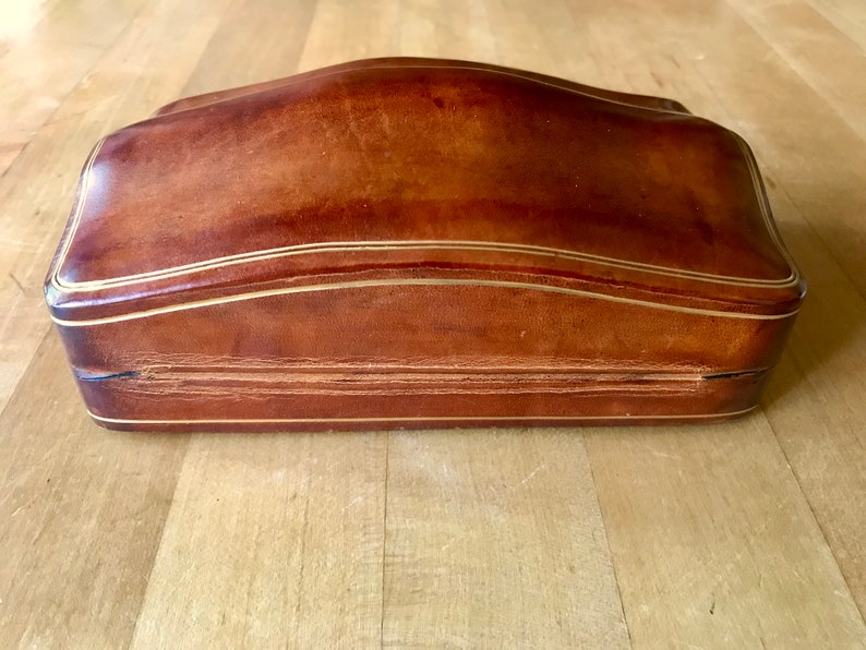 May include: A brown leather box with a gold trim. The box has a curved top and a hinged lid.