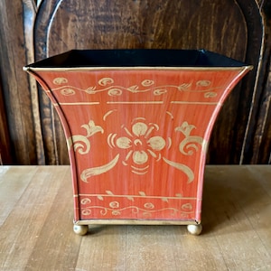 May include: A square terracotta planter with gold painted floral designs and a black rim. The planter has three gold feet.