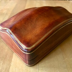 May include: A brown leather box with a gold trim. The box has a curved top and a rectangular base.