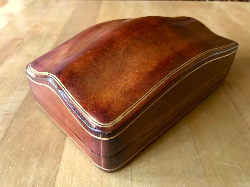 May include: A brown leather box with a gold trim. The box has a curved top and a rectangular base.