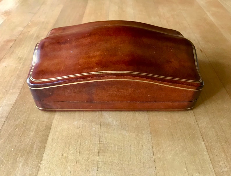 May include: A brown leather box with a curved top and gold trim. The box is closed.