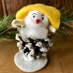 May include: A small, white, felt snowman ornament with a yellow felt hat and a red pom-pom nose. The snowman is sitting on a pine cone base with a white, snowy finish.
