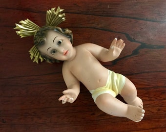 May include: A porcelain figurine of a baby Jesus wearing a yellow diaper and a gold halo. The figurine is lying on its back with its arms outstretched.