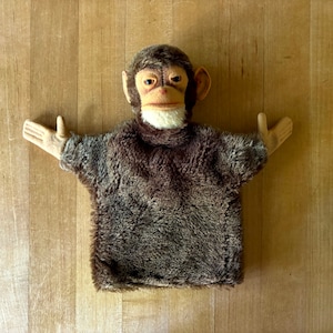 May include: A brown furry monkey hand puppet with a white face and black eyes. The puppet has a wide mouth and is smiling.