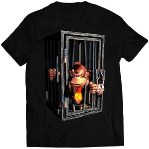 Puede incluir: Camiseta negra con un gráfico de Donkey Kong tras las rejas. El simio de dibujos animados lleva una corbata roja con "DK". La puerta de la jaula está cerrada con un candado. El diseño está centrado en la camiseta.