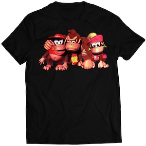 Puede incluir: Una camiseta negra con un gráfico de Donkey Kong, Diddy Kong y Dixie Kong. Los Kongs llevan sus atuendos característicos de la serie de videojuegos Donkey Kong Country.