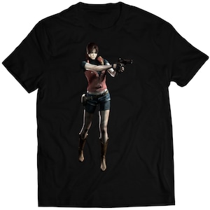 May include: Black t-shirt featuring a female character from a video game holding a gun. She is wearing a red vest, blue shorts, and brown boots.