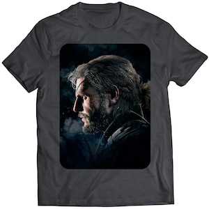 May include: Dark grey t-shirt with a portrait of a man in profile, sporting long hair, a beard, and an eye patch. The image is set against a dark, smoky backdrop. The man has a scar across his face.