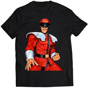 Könnte beinhalten: Schwarzes T-Shirt mit einer bunten Illustration eines Videospielcharakters, der eine rot-weiße Uniform und eine rote Mütze trägt. Der Charakter hat einen ernsten Gesichtsausdruck und hält seine Faust hoch.