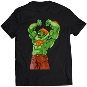 Könnte beinhalten: Schwarzes T-Shirt mit einer grünen und orangen Illustration eines muskulösen Charakters mit einem wütenden Ausdruck. Der Charakter beugt beide Arme und hat einen roten Gürtel.