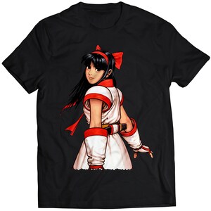 Könnte beinhalten: Schwarzes T-Shirt mit einer bunten Illustration einer Frau mit langen schwarzen Haaren, die ein weiß-rotes Outfit trägt. Sie schaut über ihre Schulter und hat ein rotes Band im Haar.