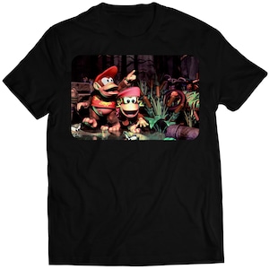 Puede incluir: Una camiseta negra con un estampado gráfico de dos monos en un entorno de selva. Los monos llevan gorras rojas y están de pie en una escena de selva verde con árboles y enredaderas.