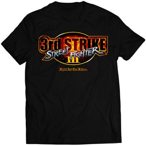 May include: Black t-shirt with a red and yellow graphic of the Street Fighter III: 3rd Strike logo. The logo features the text "3rd Strike" and "Street Fighter III" in a stylized font. The text "Fight for the Future" is below the logo.